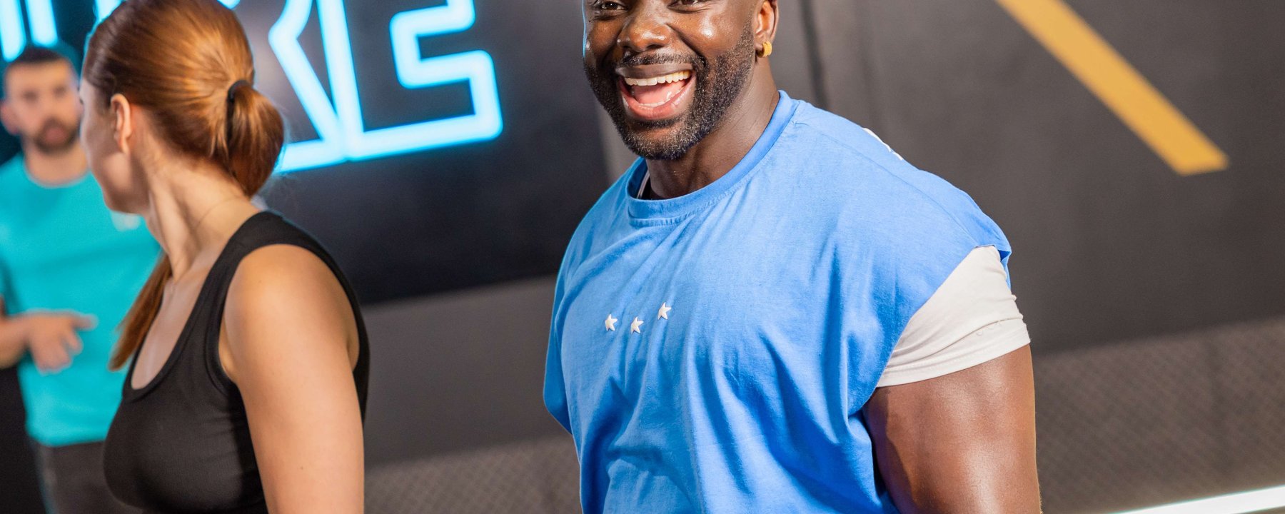 Smiling man in blue sleeveless shirt with two people in the background at gym