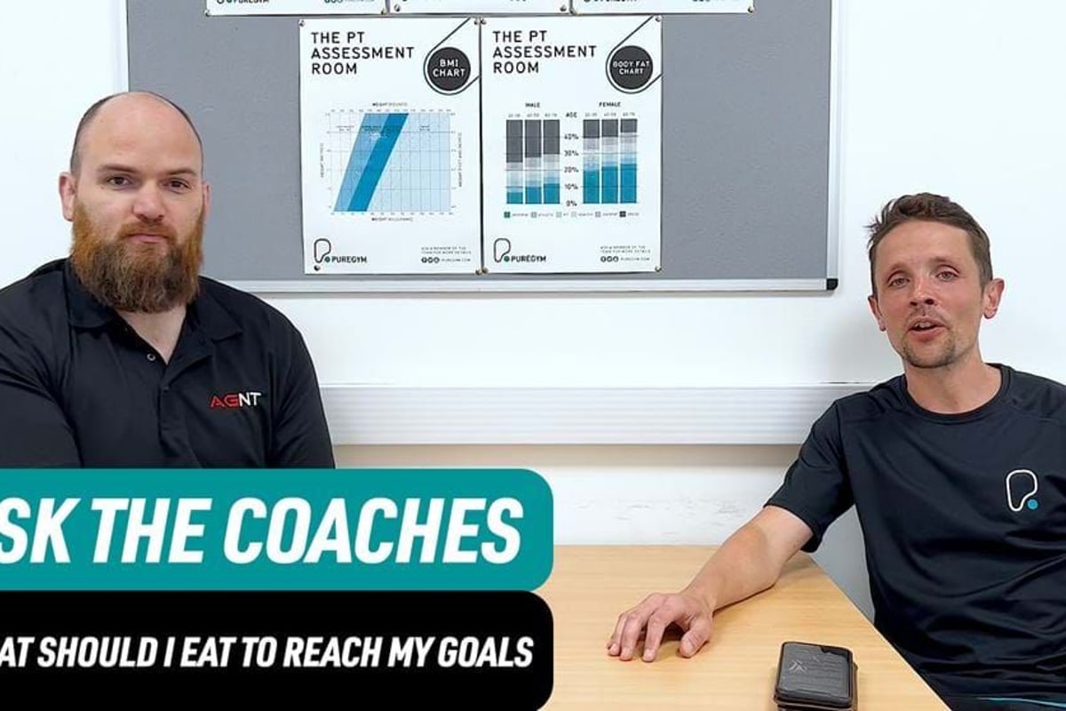 Two men sitting at a table with fitness charts on the wall behind them.
