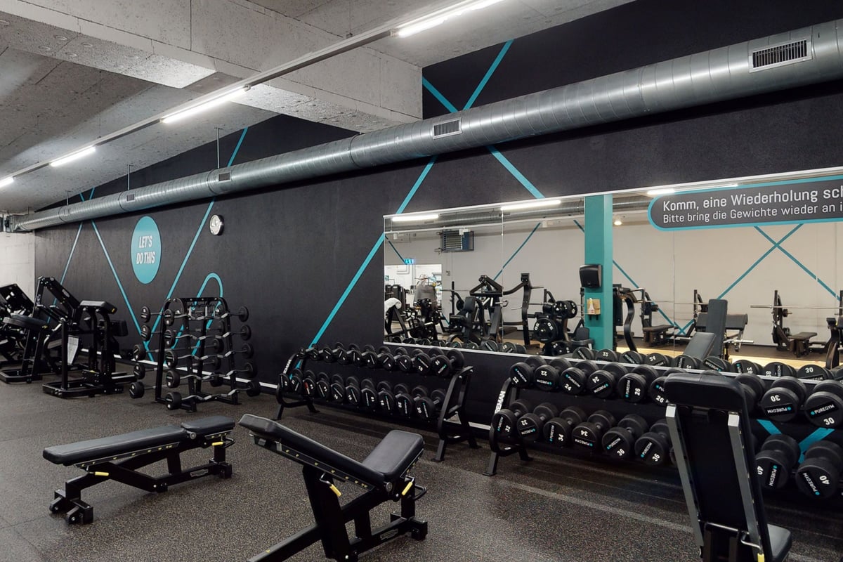 PureGym Pfäffikon: Kraftraum mit Hantelablagen, Bänken und Lüftungsrohren an der Decke mit blaugrünen Akzenten an dunklen Wänden.