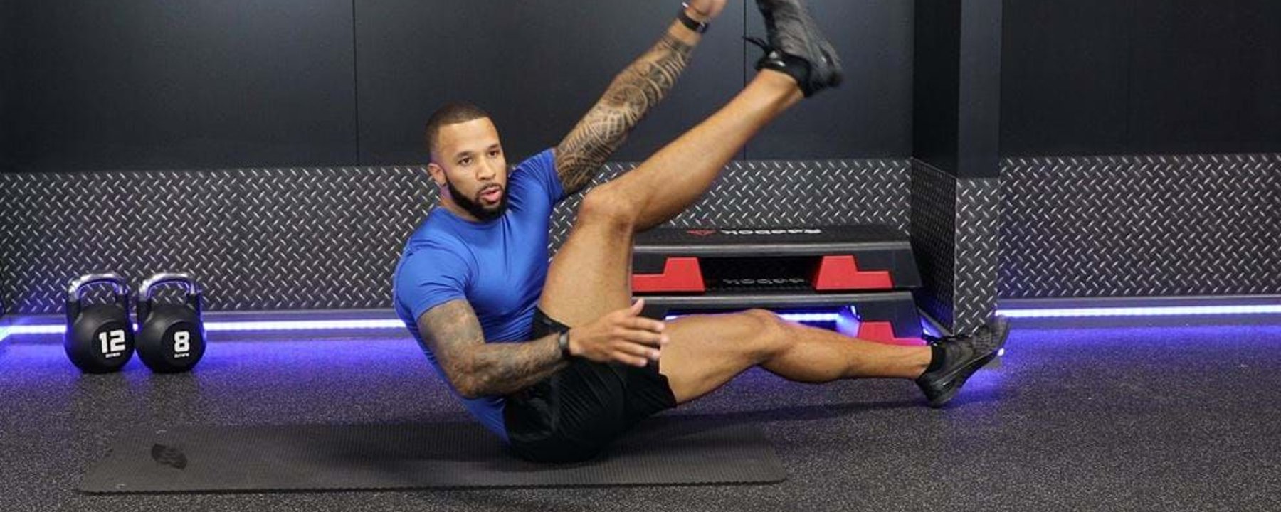Man in blue shirt doing a seated leg raise exercise on a gym mat