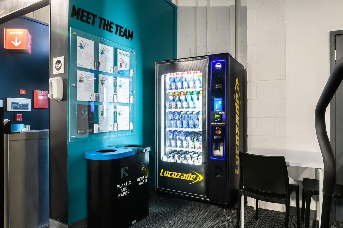 PureGym Buckingham: Beverage vending machine next to personal trainer profiles in an informational wall display.