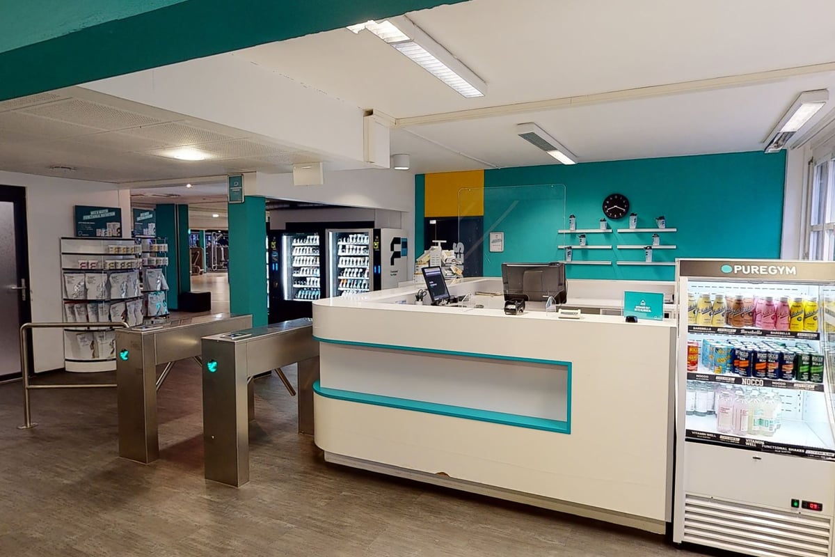 PureGym Zürich Lochergut: Intérieur de l'aire de réception d'une salle de sport avec distributeurs automatiques, kiosques en libre-service et comptoir de service.