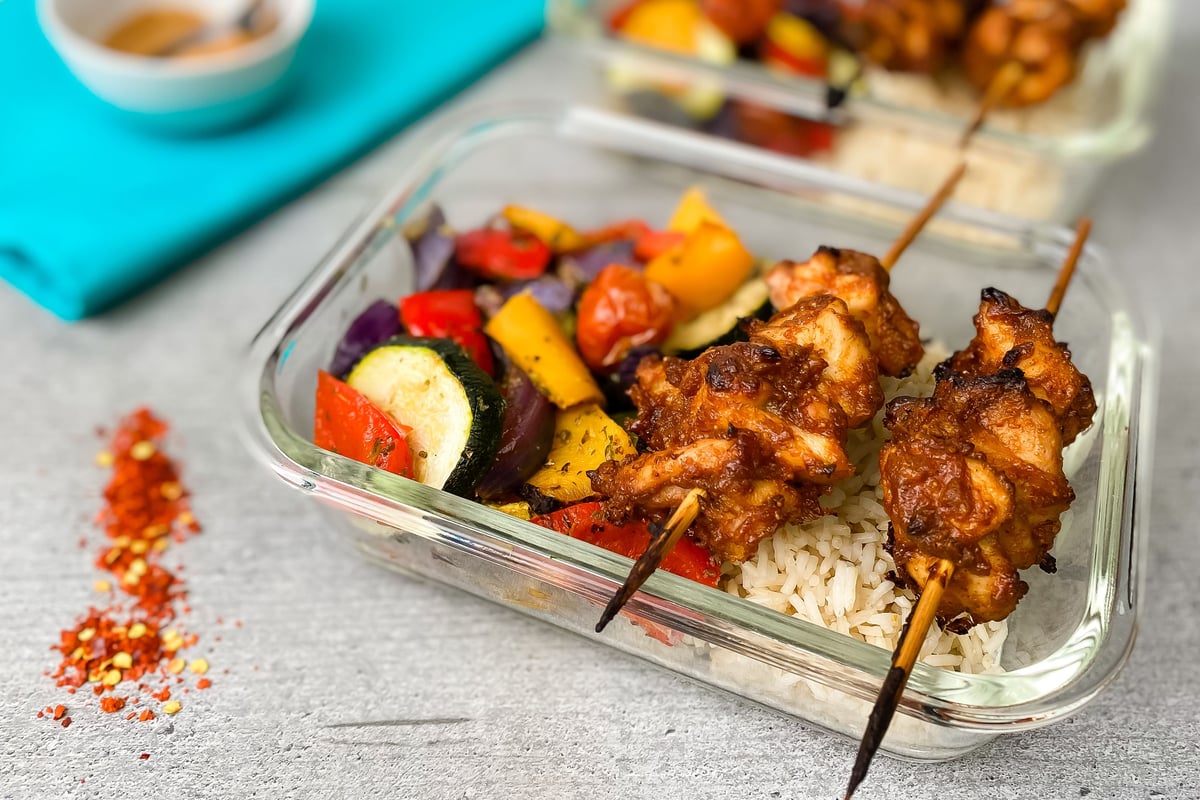 Grilled chicken skewers on rice with roasted vegetables in clear container