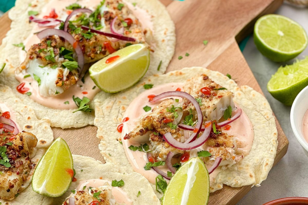 Crispy fish tacos with lime wedges, red onion and sauce on wooden board