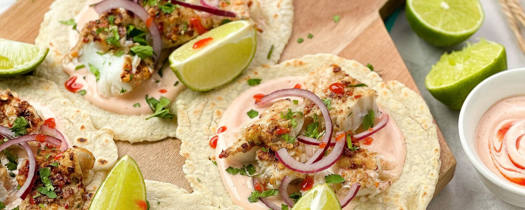 Crispy fish tacos with lime wedges, red onion and sauce on wooden board