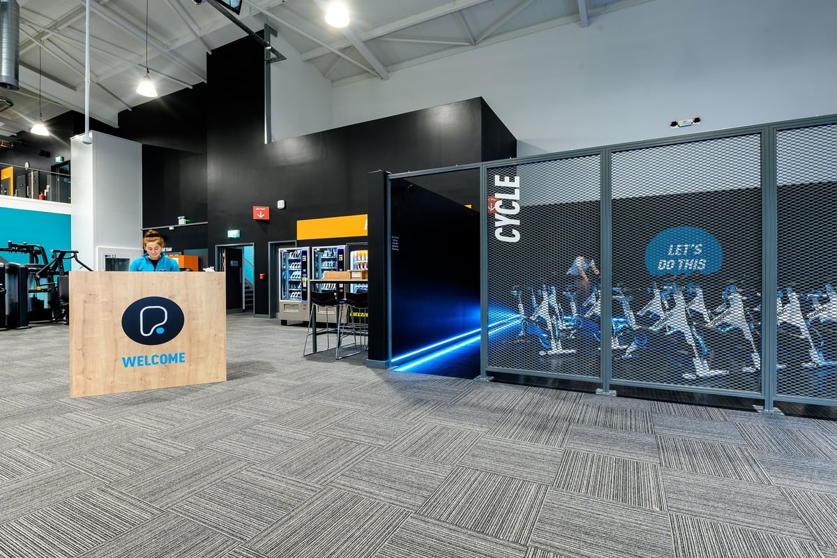 PureGym Exeter Bishops Court: Welcome desk, spin cycle studio area with bikes, dark colour scheme, unattended kiosks.