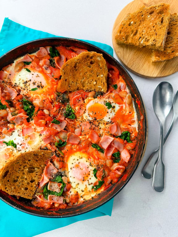 Shakshuka with eggs, spinach, ham, and toasted bread in a pan with two spoons nearby
