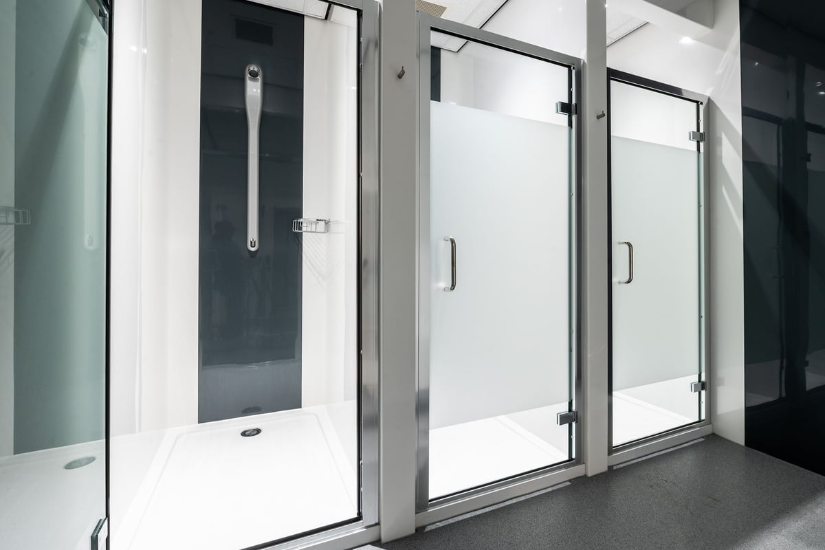 PureGym Newbury: Rows of upright shower stalls with glass doors and tiled flooring.