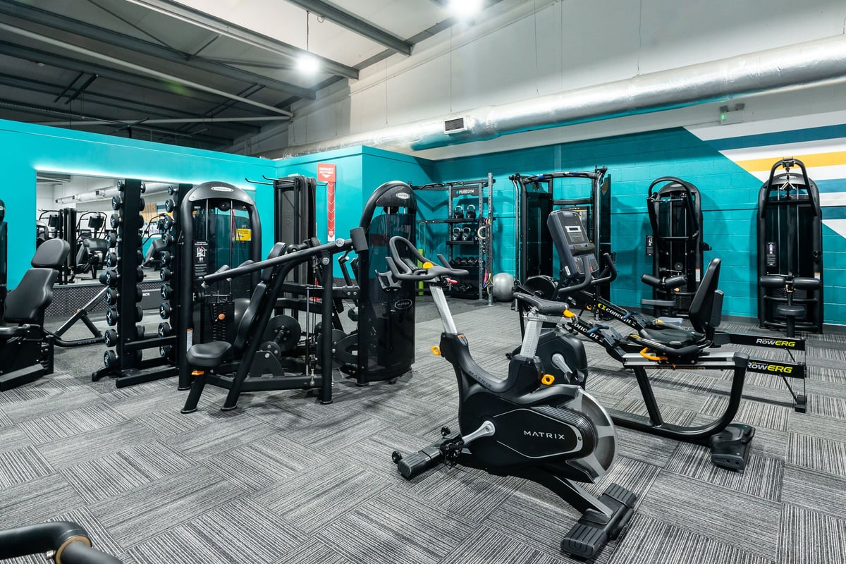 PureGym Wrexham: Gym studio with weight machines, exercise bikes, benches, and teal accent walls.