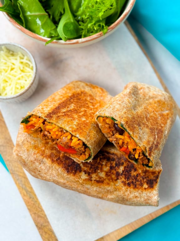 Grilled burrito cut in half showing rice, beans, and vegetables with salad and cheese bowl