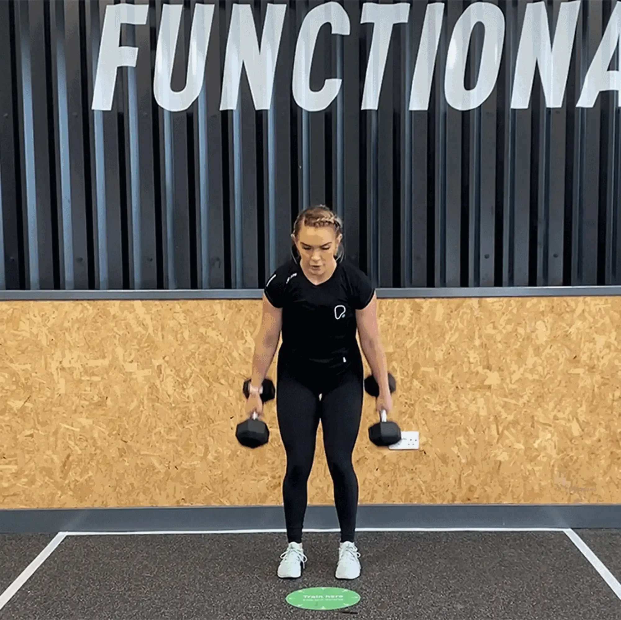 Woman in black workout clothes lifting dumbbells in gym with FUNCTIONAL sign above