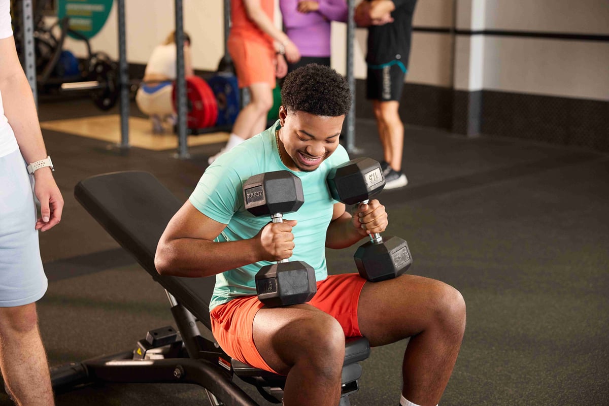 Man sitting on bench holding dumbbells in gym with people in background