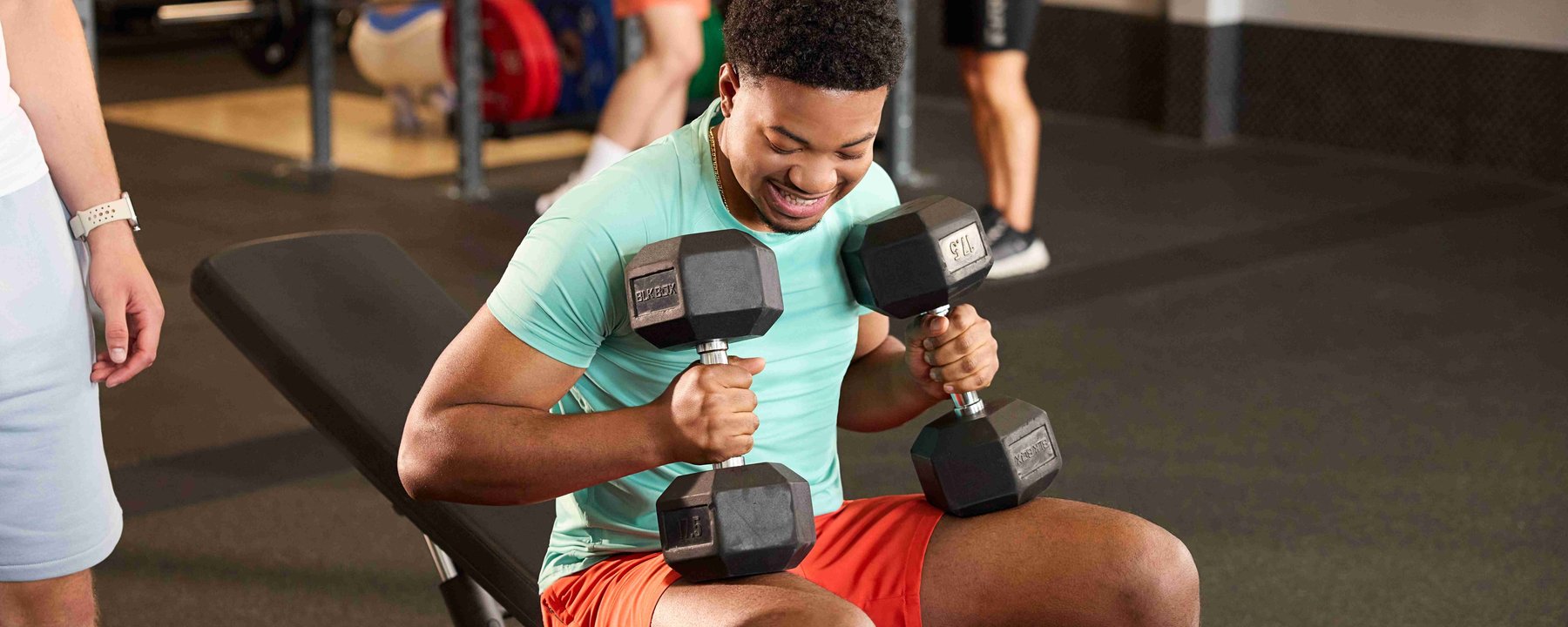 Man sitting on bench holding dumbbells in gym with people in background
