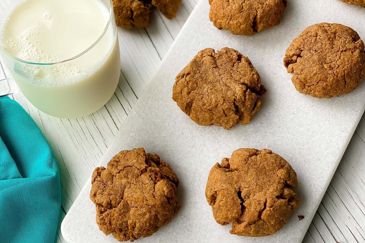 Six brown cookies on a white board with a glass of milk and scattered cocoa nibs nearby
