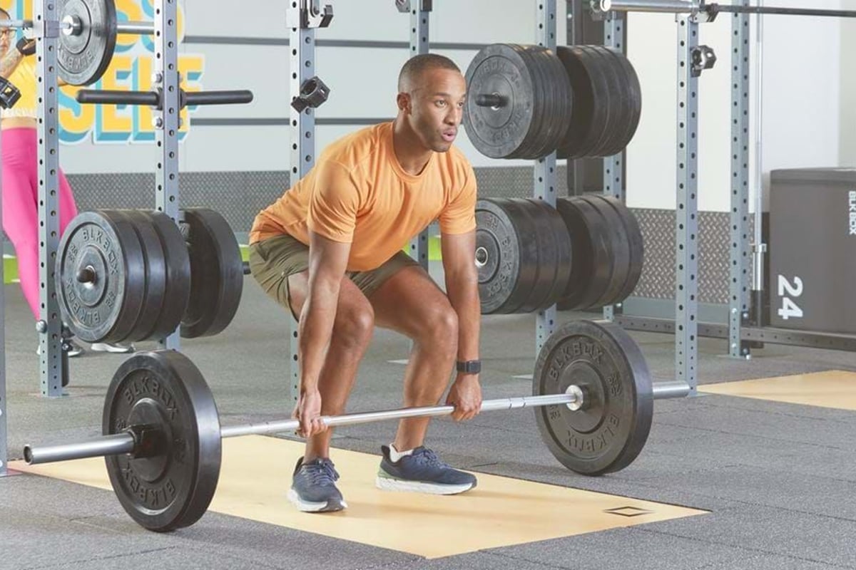 Man in orange shirt lifting barbell in gym with weight plates on racks behind him