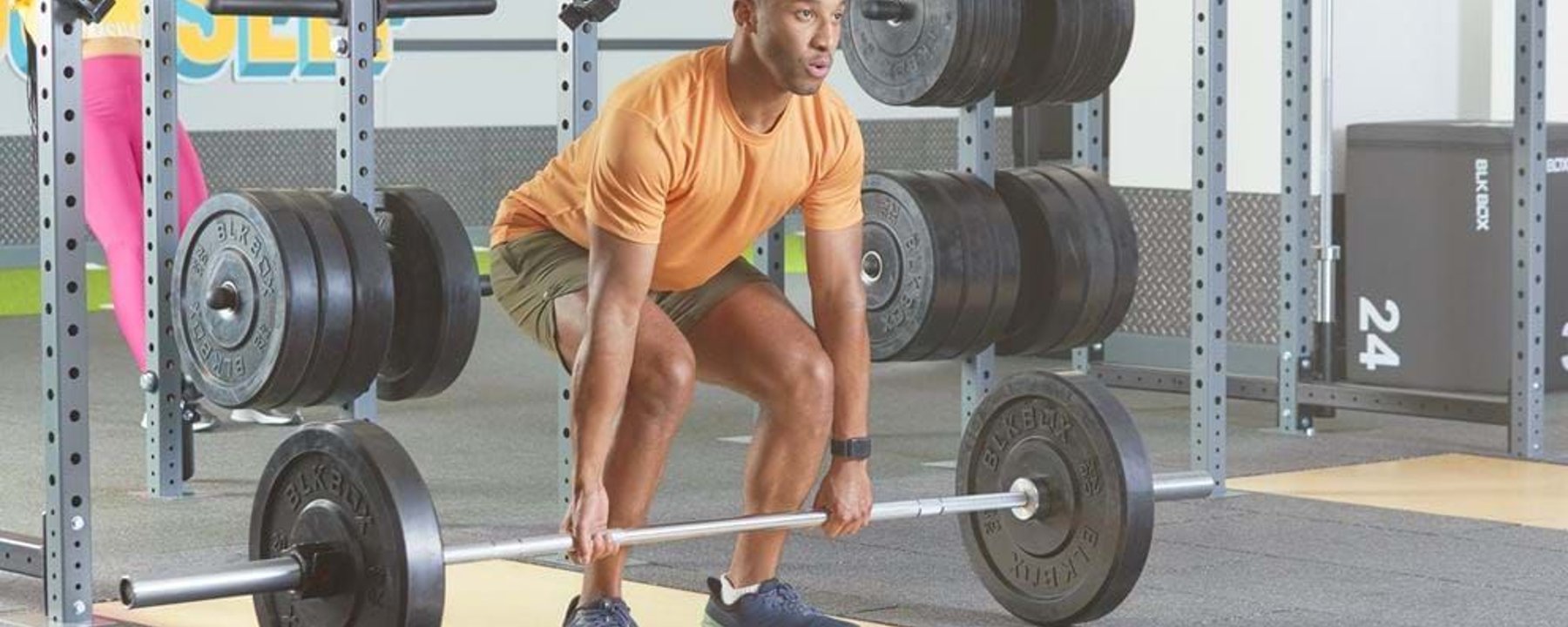 Man in orange shirt lifting barbell in gym with weight plates on racks behind him