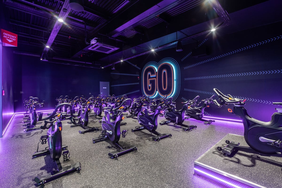 PureGym Manchester Cheetham Hill Retail Park: Rows of stationary bikes in a purple-lit studio with neon 'GO' signage.