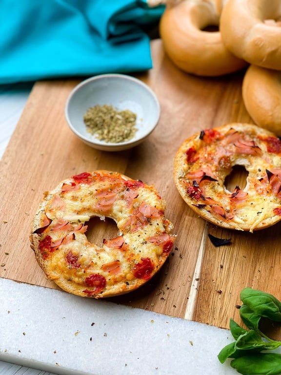 Two toasted bagel halves with melted cheese and ham on a wooden board with herbs nearby