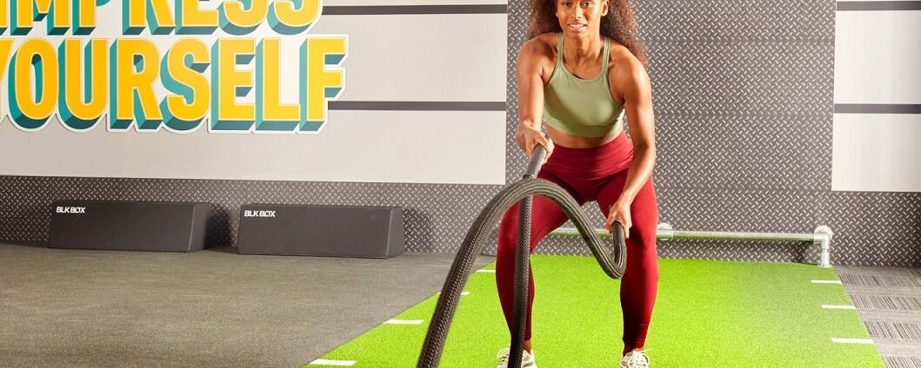 Woman exercising with battle ropes on green turf in gym with Impress Yourself sign