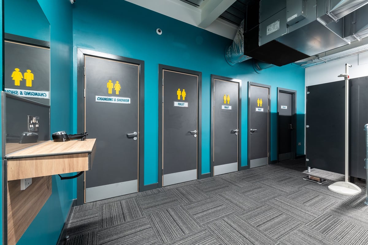 PureGym Droitwich Spa: Rows of entry gates with teal accents, sink counter, and patterned floor tiling.