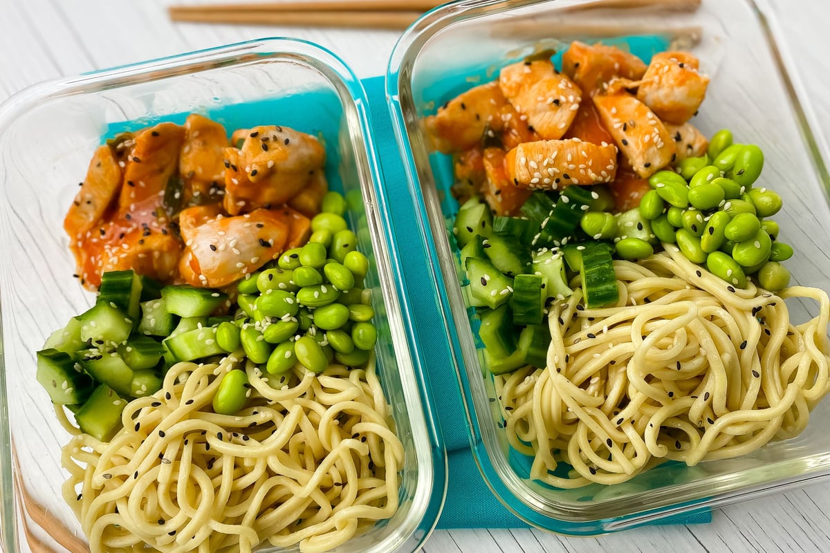 Two meal prep containers with noodles, edamame, cucumber and sesame chicken pieces