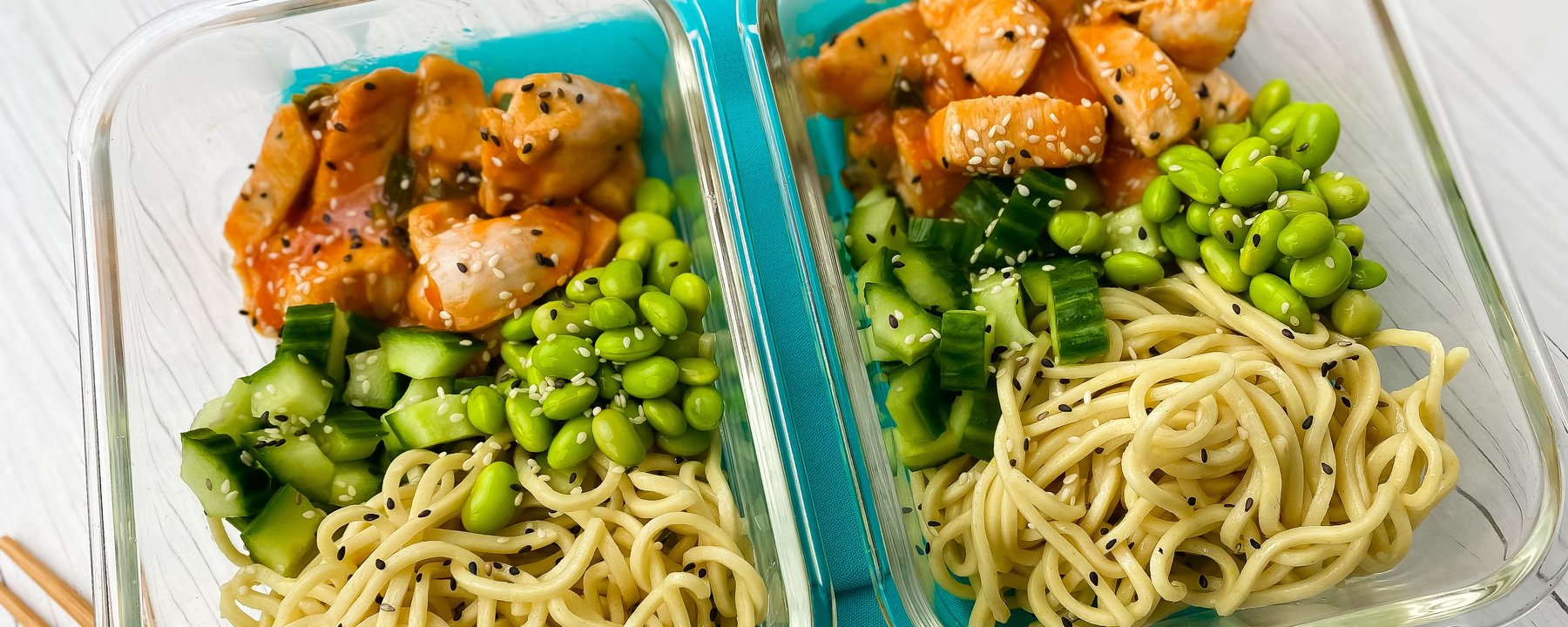 Two meal prep containers with noodles, edamame, cucumber and sesame chicken pieces