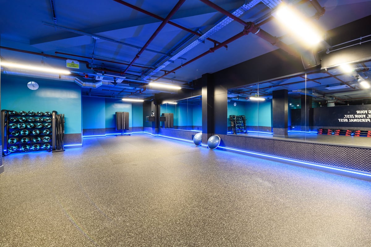 PureGym Basildon: Exercise studio with wall mirrors, weight racks, medicine balls, rubberized flooring under blue lighting.