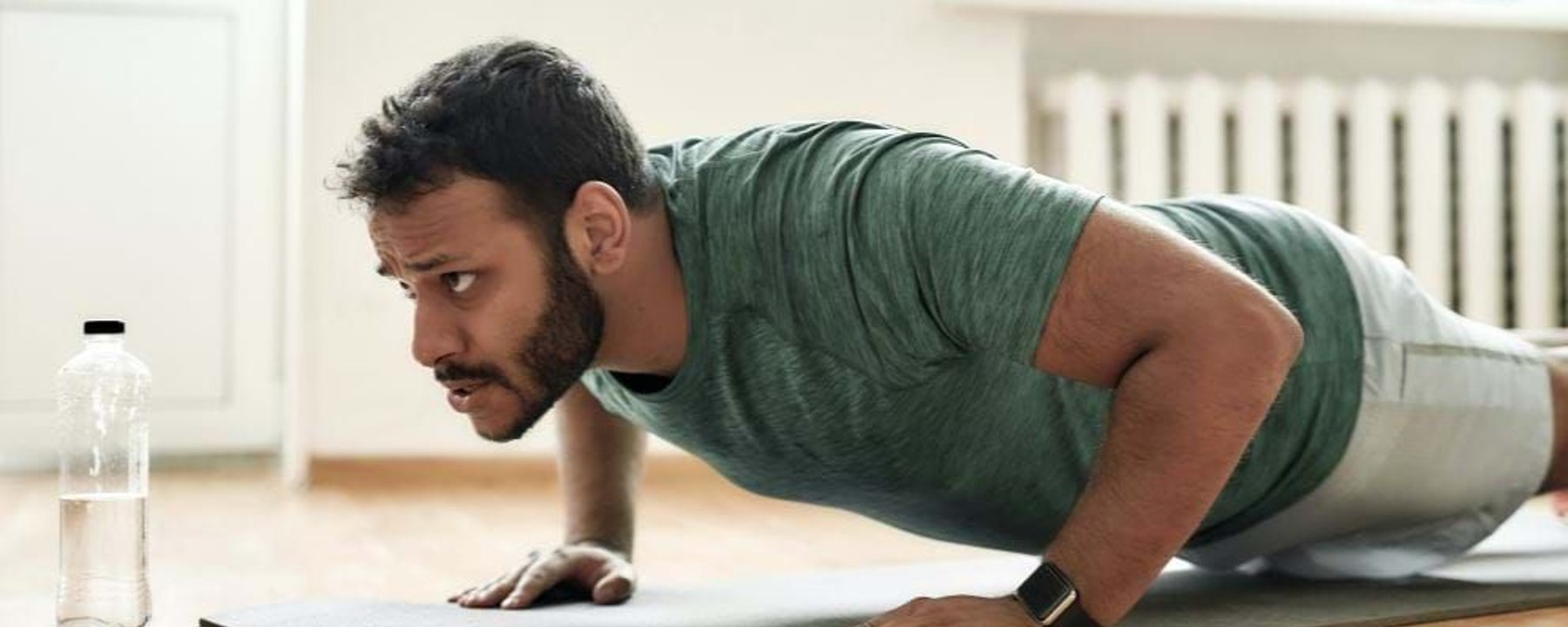 Man in green shirt doing push-ups on a mat with a water bottle nearby indoors
