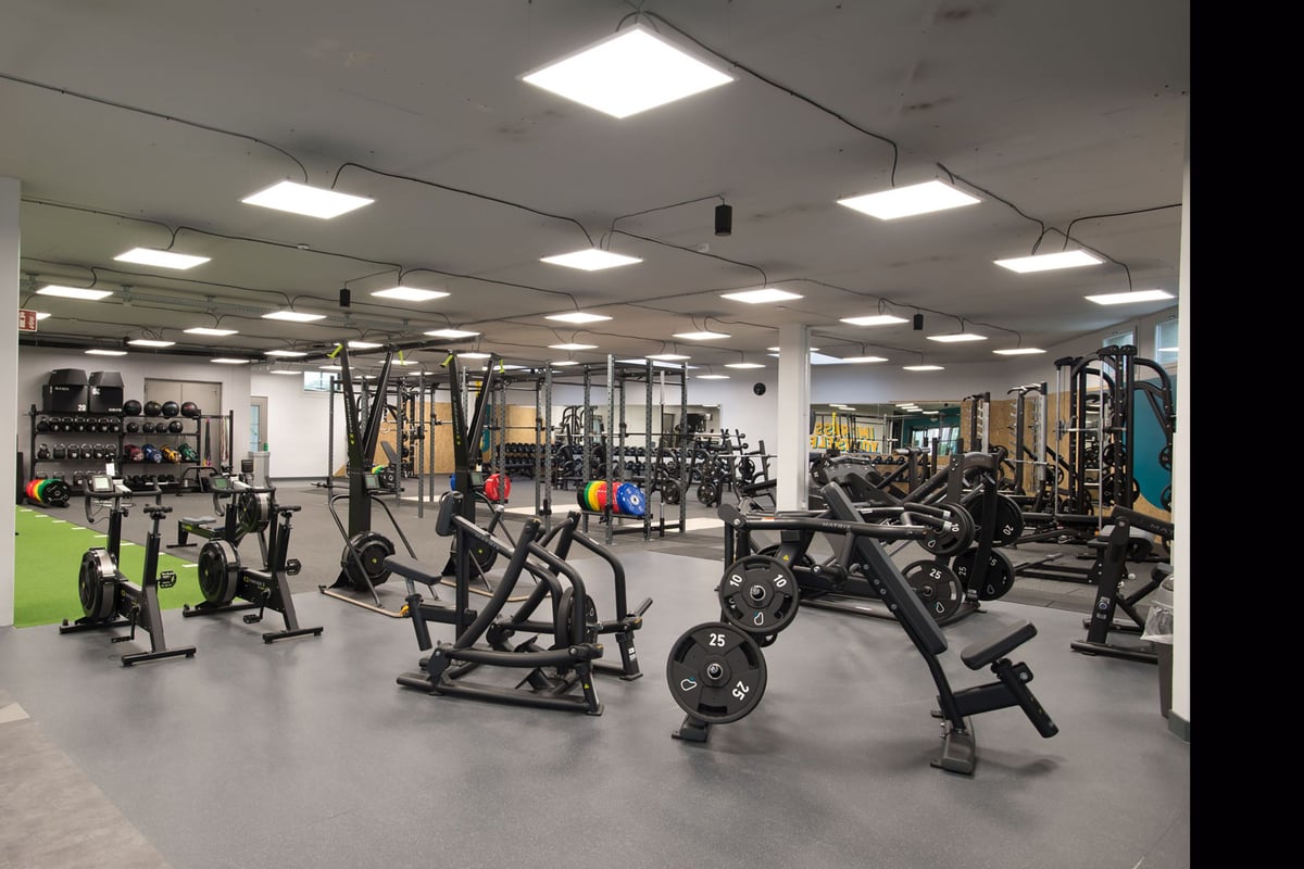 PureGym Lyss: Salle de sport avec appareils de musculation, bancs, haltères, supports et sols caoutchoutés sous un éclairage intense.