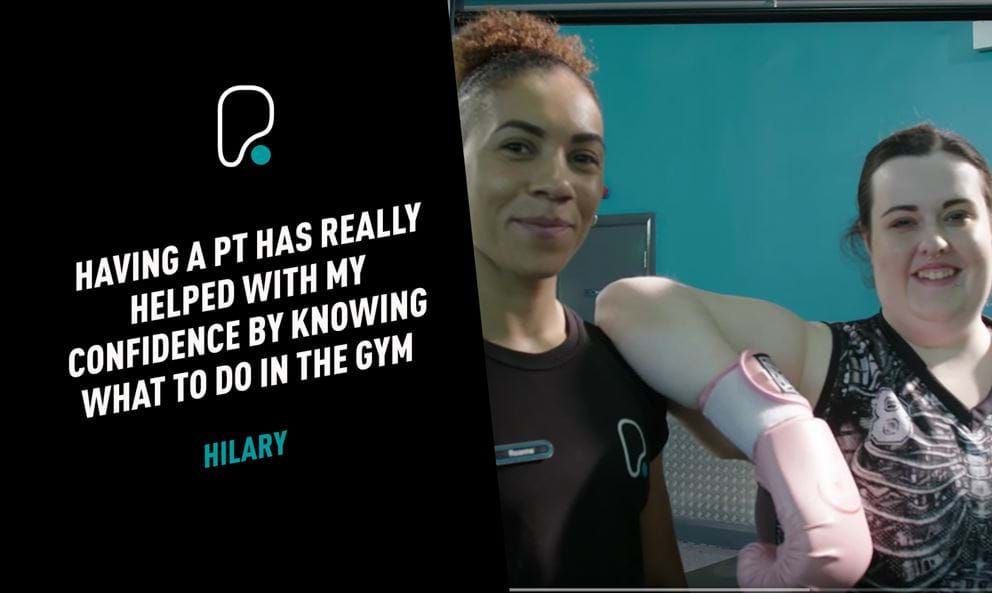 Two women smiling in a gym, one wearing pink boxing gloves and sportswear.