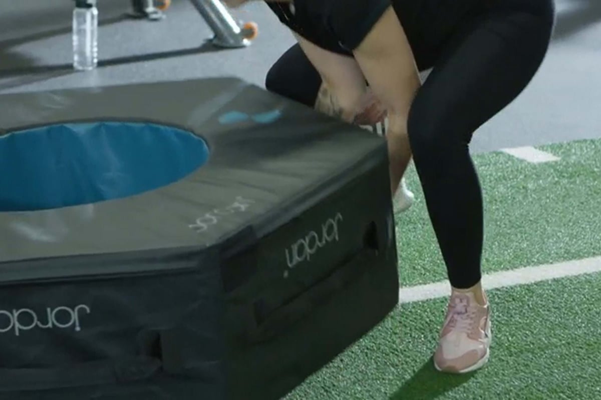 Person in black leggings and pink trainers lifting a padded exercise box on turf flooring