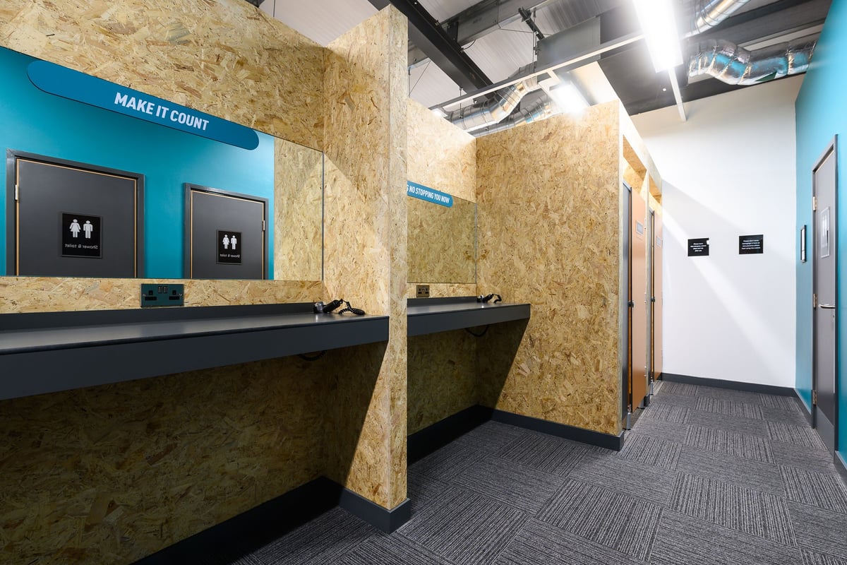 PureGym Barnstaple: Reception area with kiosks, benches, wood-paneled walls, and directional signage reading "Make It Court".