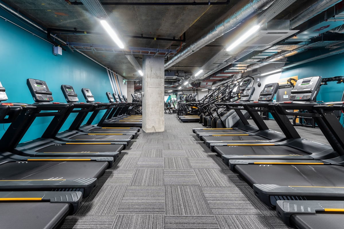 PureGym London Hoxton: Rows of modern treadmills in a dimly lit gym interior with teal walls and concrete pillars.