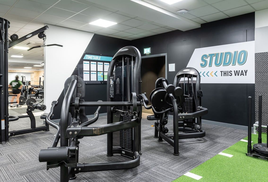 PureGym Epsom: Weight machines, exercise studio signage, black and grey colour scheme, turf floor area.