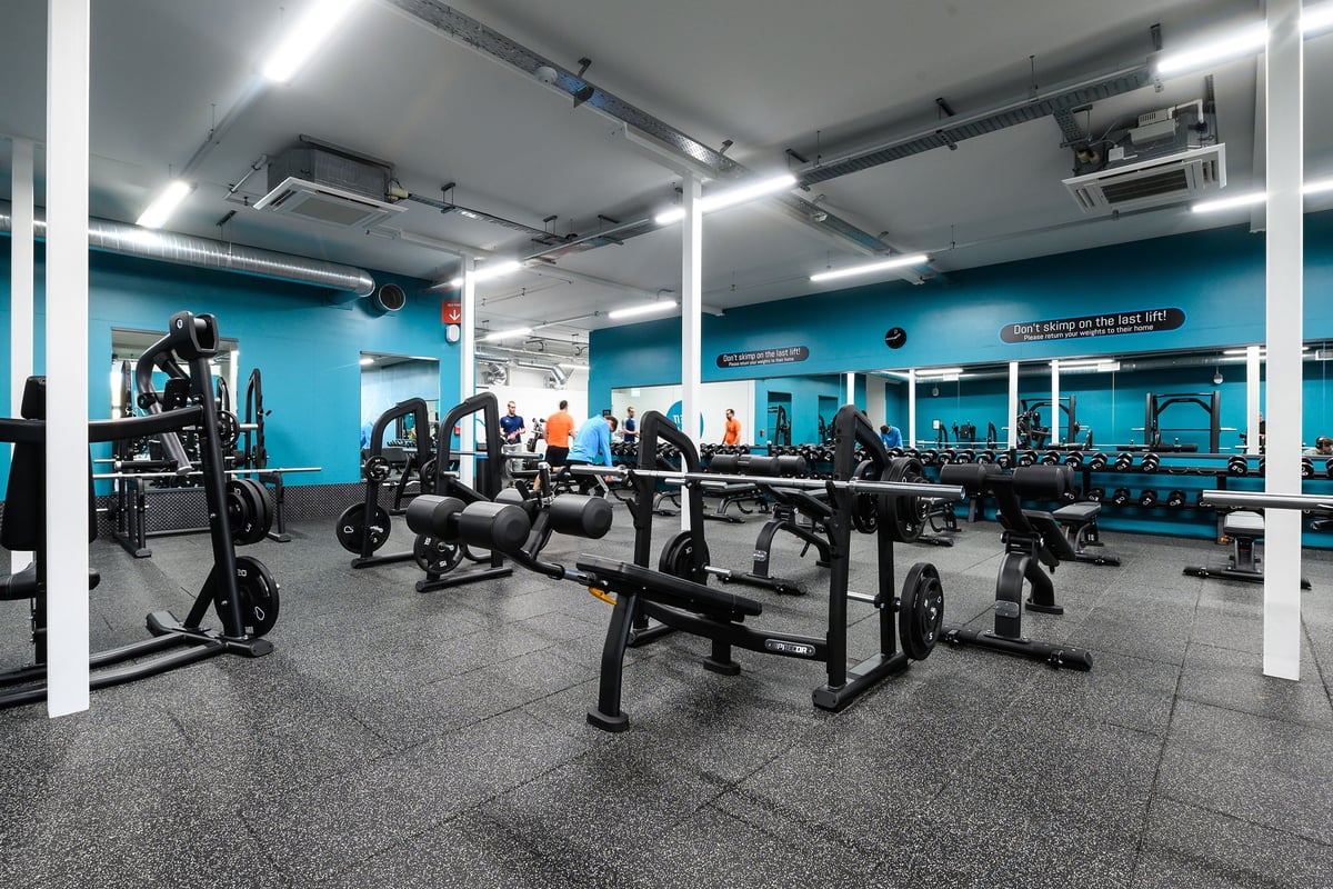 PureGym Exeter Bishops Court: Rows of weight benches, freeweights, mirrors, and rubberized flooring in a teal-coloured gym.