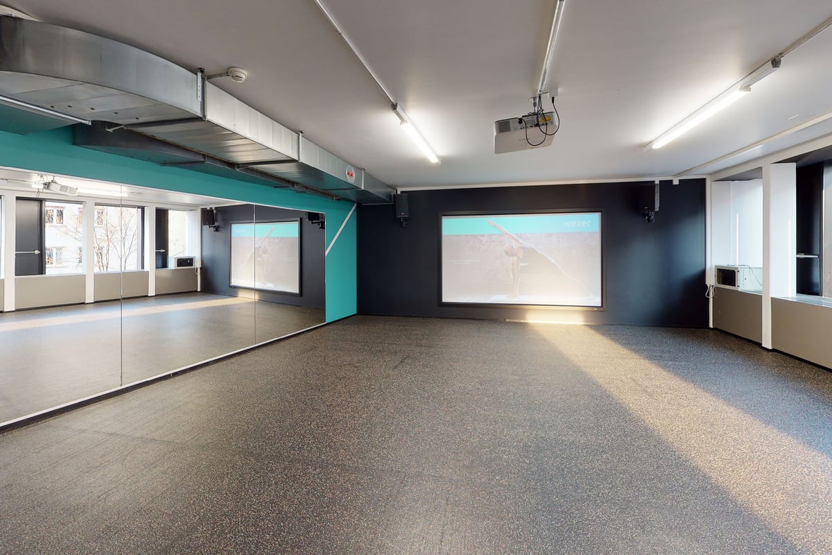 PureGym Basel Clara Huus: Grosser leerer Raum mit blaugrünen Akzenten, gummiertem Boden, Wandschirmen und verspiegelten Wänden.