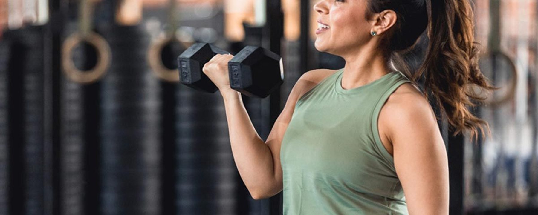 Femme en haut vert soulevant un haltère en salle avec poids empilés derrière