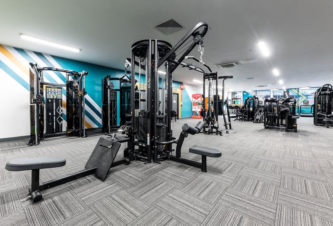 PureGym Bangor Wales: Gym studio with weight machines, benches, mirrors along walls, and rubberized patterned flooring.