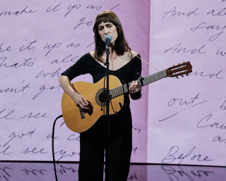 Leslie Feist is wearing black and playing an acoustic guitar against a pink background