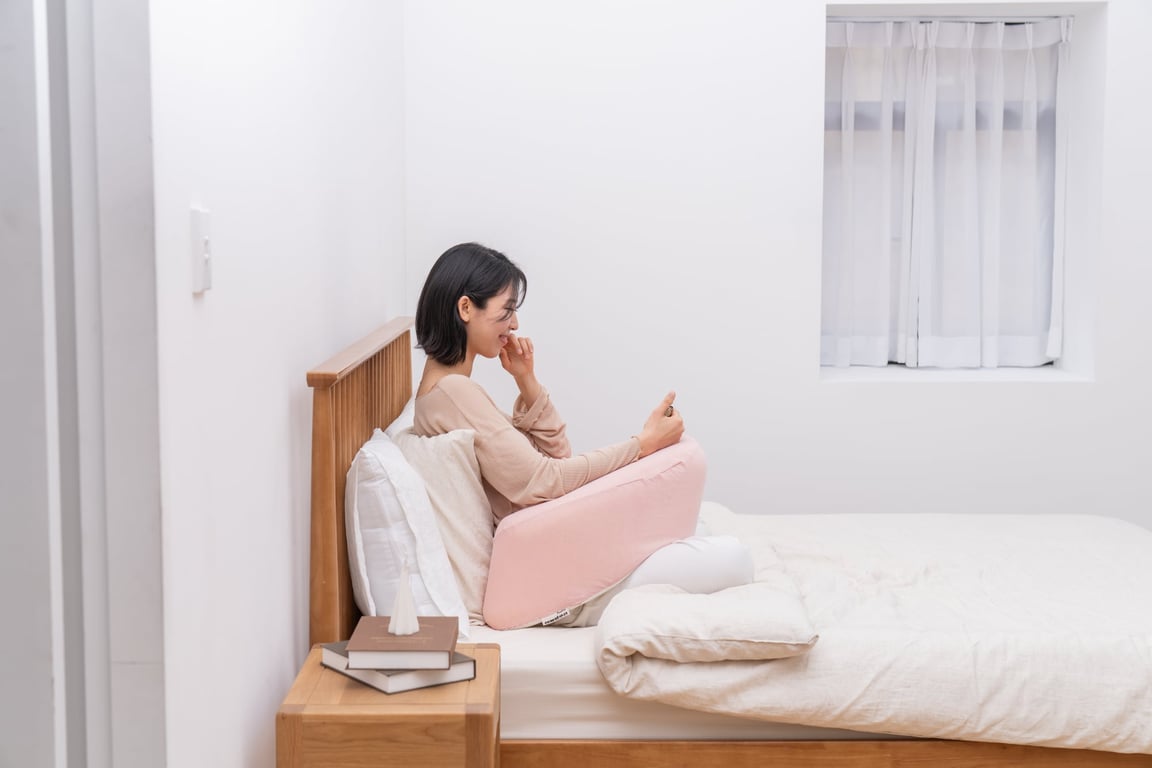 스마트폰을 보며 웃고 있는 단발머리의 30대 여성이 베드헤드에 기대어 앉아, 허리와 팔 아래에 분홍색 레벨업쿠션을 넣어 편안한 자세로 휴식을 취하고 있습니다.