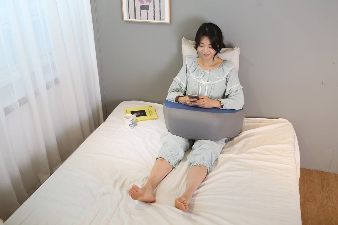 20대 후반~30대 초반 한국 여성이 밝은 침실 침대 위에 등받이 쿠션에 기대어 앉아, 회색·네이비 투톤의 레벨업쿠션을 무릎 위에 올리고 스마트폰을 사용하고 있는 편안한 홈웨어 모습.