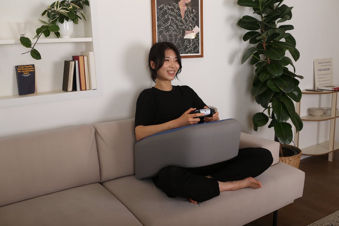 소파 위에서 PS5 컨트롤러를 잡고 게임을 즐기는 여성의 무릎 위에 놓인 그레이 레벨업쿠션