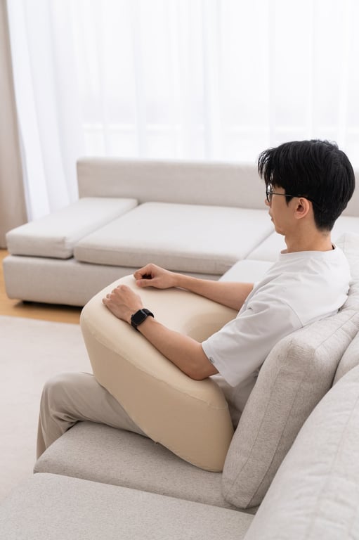 소파에서 편안하게 휴식하며 베이지색 펌플렉스 레벨업쿠션에 팔을 resting하는 남성으로, 팔과 어깨 완화를 위한 제품의 지지력을 강조