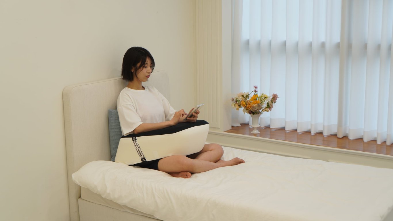 침대 위에 앉아 스마트폰을 보며 팔을 쿠션 위에 편안하게 올린 여성의 모습
