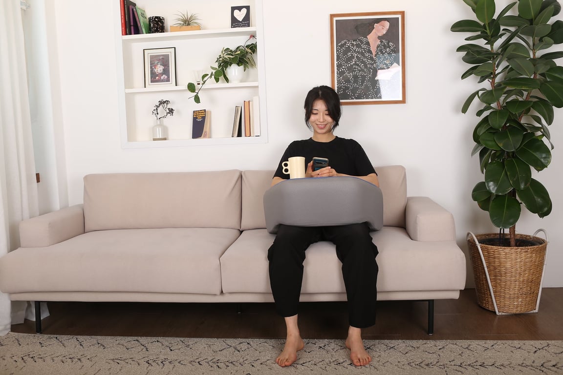 밝은 거실 소파에 앉은 여성이 레벨업쿠션을 무릎 위에 올려 팔을 받치며 스마트폰을 편안하게 사용하는 모습