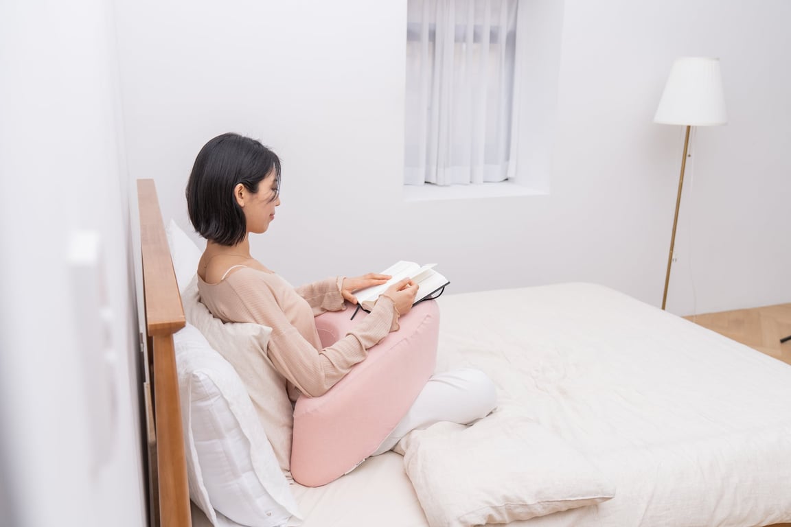 침대 위에서 책을 읽으며 베이지색 펌플렉스 레벨업쿠션에 팔을 resting하고 있는 여성으로, 쿠션이 팔 무게를 지지하여 어깨 strain을 완화하는 방법을 보여줌