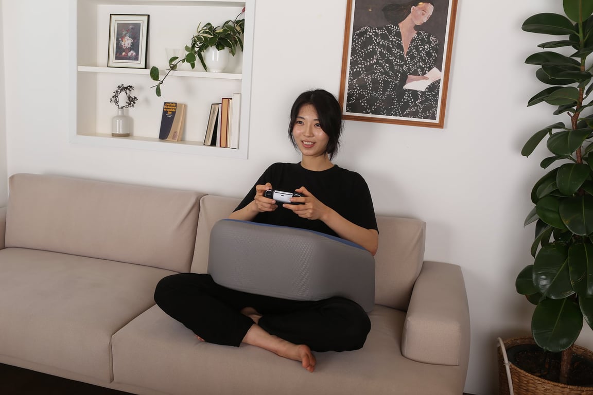 ソファに座った女性がレベルアップクッションを膝の上に置き、ゲームコントローラーを両手で持ってゲームをプレイしている様子