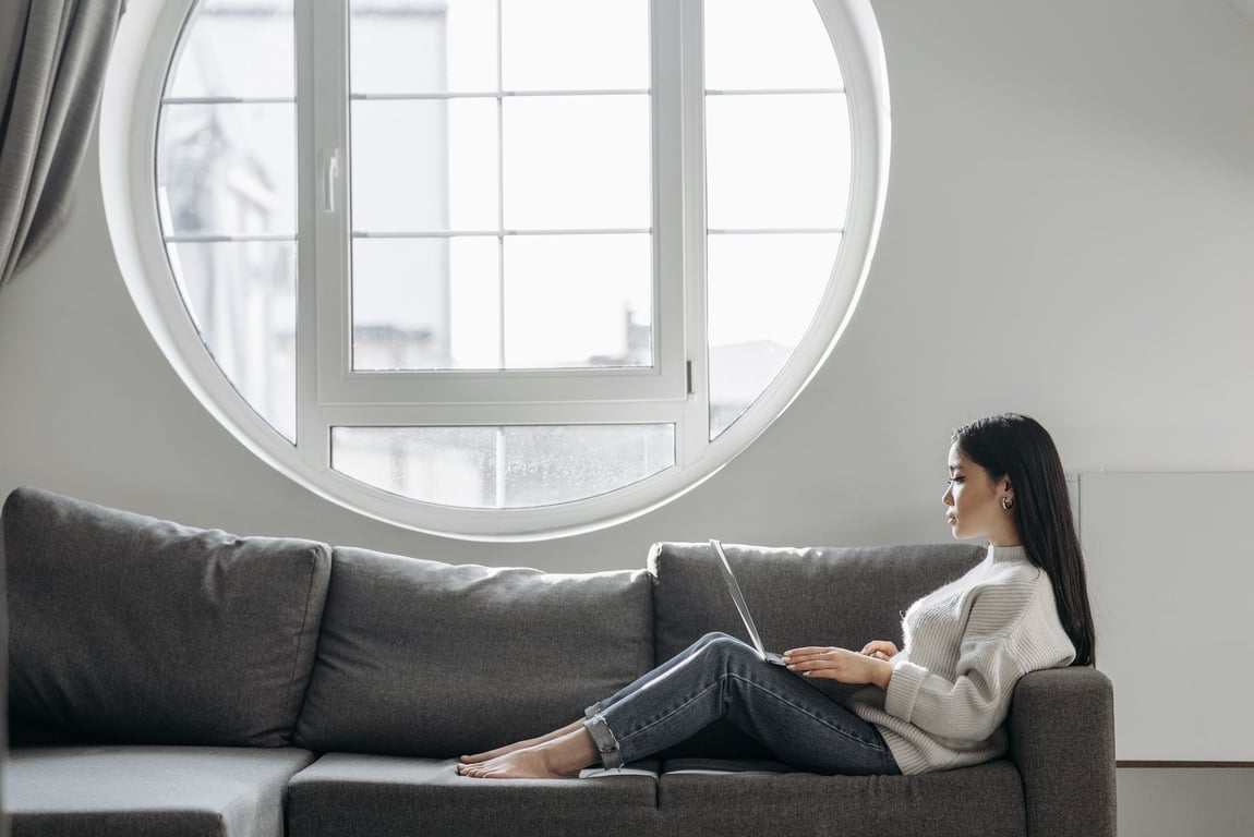 A woman rests on a sofa using a laptop, with natural light streaming through a round window.