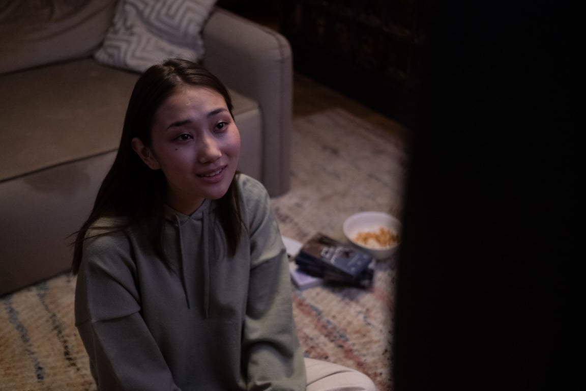 夜の薄暗いリビングで床に座り、テレビを見ながらお菓子を食べる若い女性