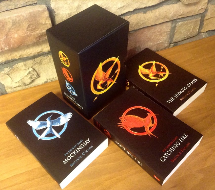 A collection of The Hunger Games book series boxes arranged on a wooden surface against a brick wall background.